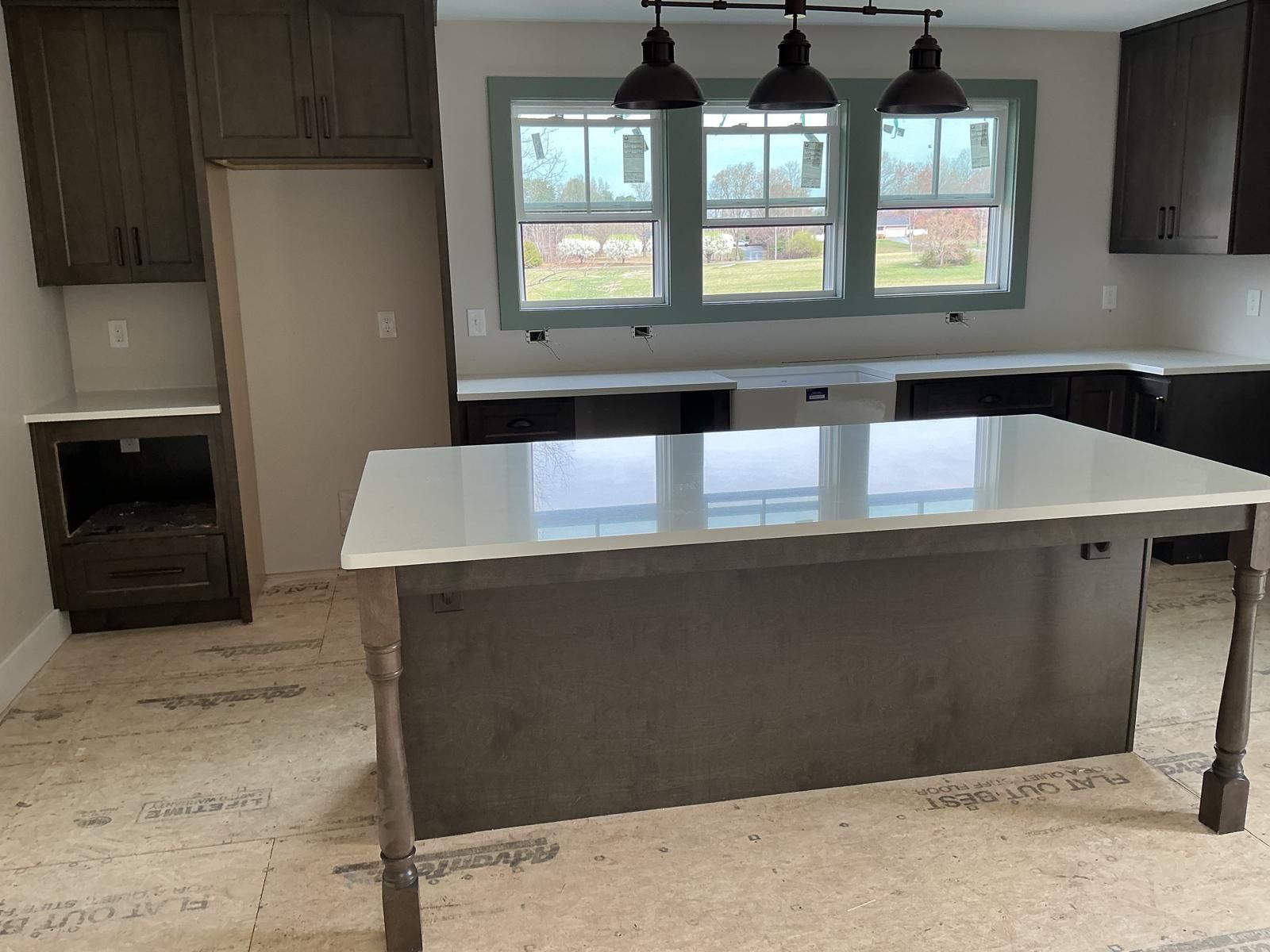 Empty modern kitchen with white island, dark cabinets, pendant lights, and three windows.