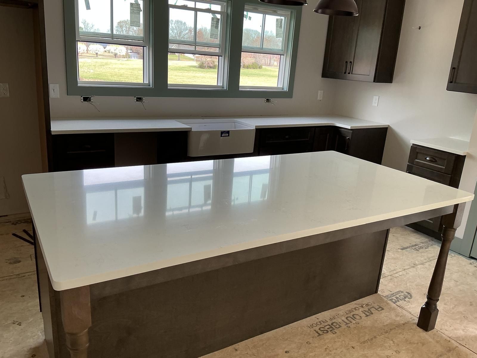 Empty kitchen with a large white island table, window above the sink, and cabinets along the walls.