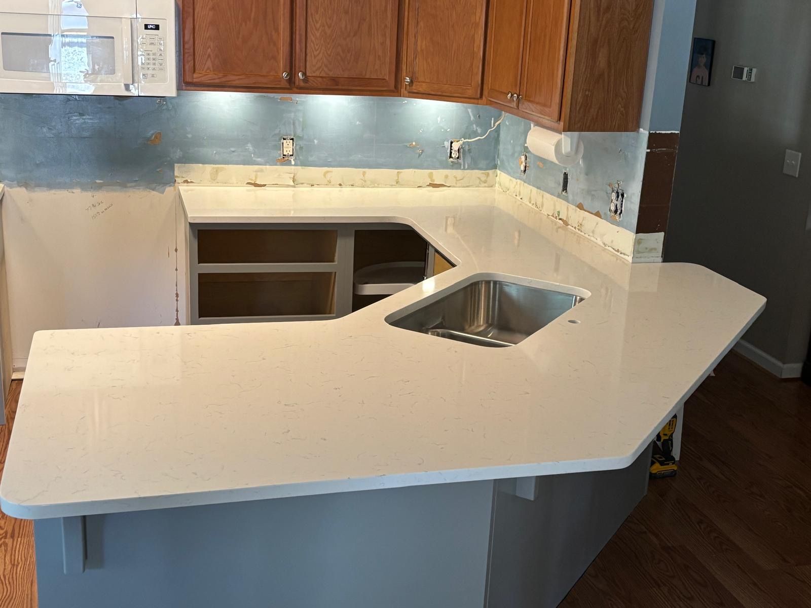 Kitchen with white countertop and stainless sink, unfinished cabinets, and a wall with gray primer and backsplash under installation