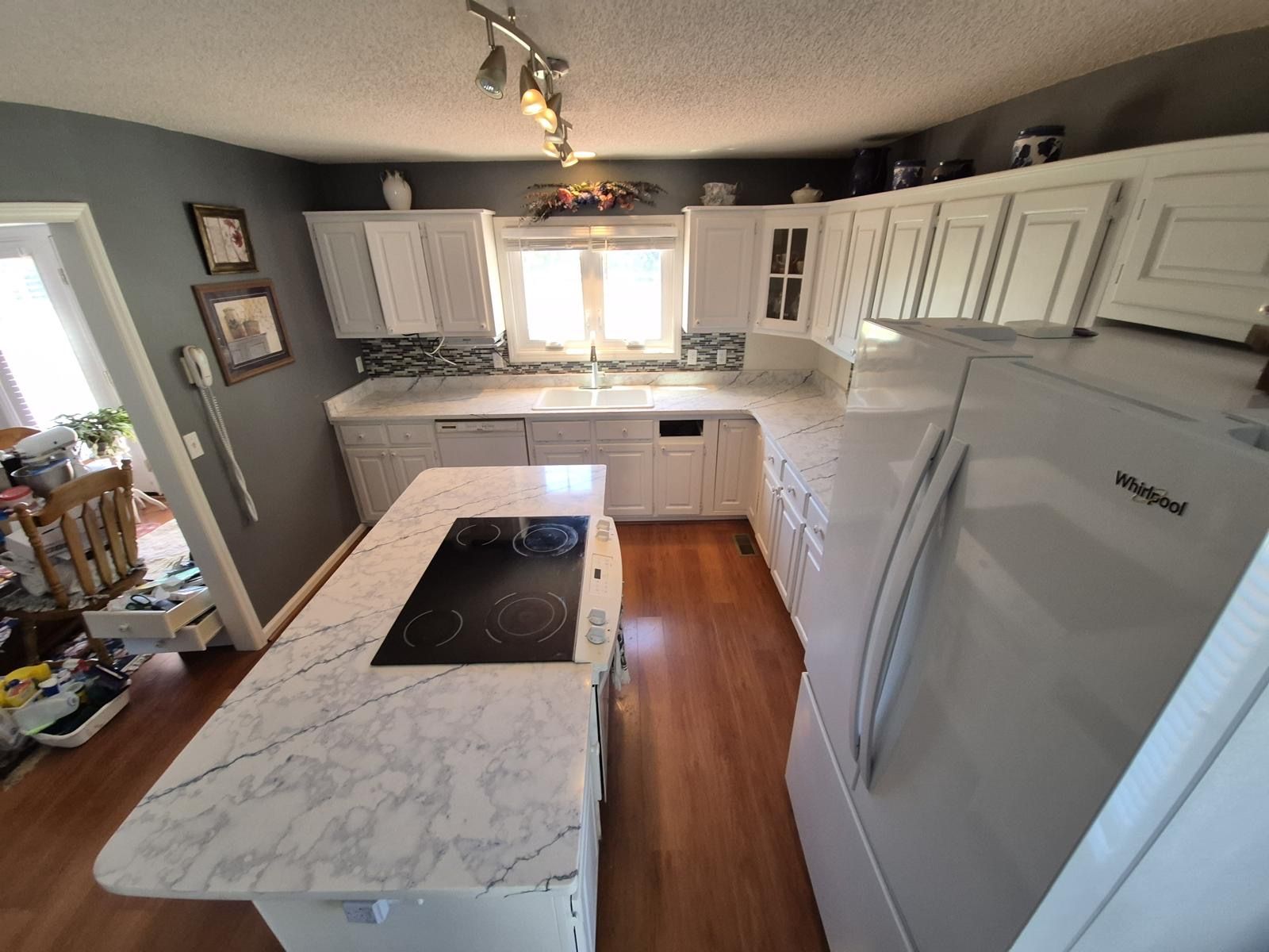 Small white kitchen with marble counters, island cooktop, gray walls, and stainless refrigerator.