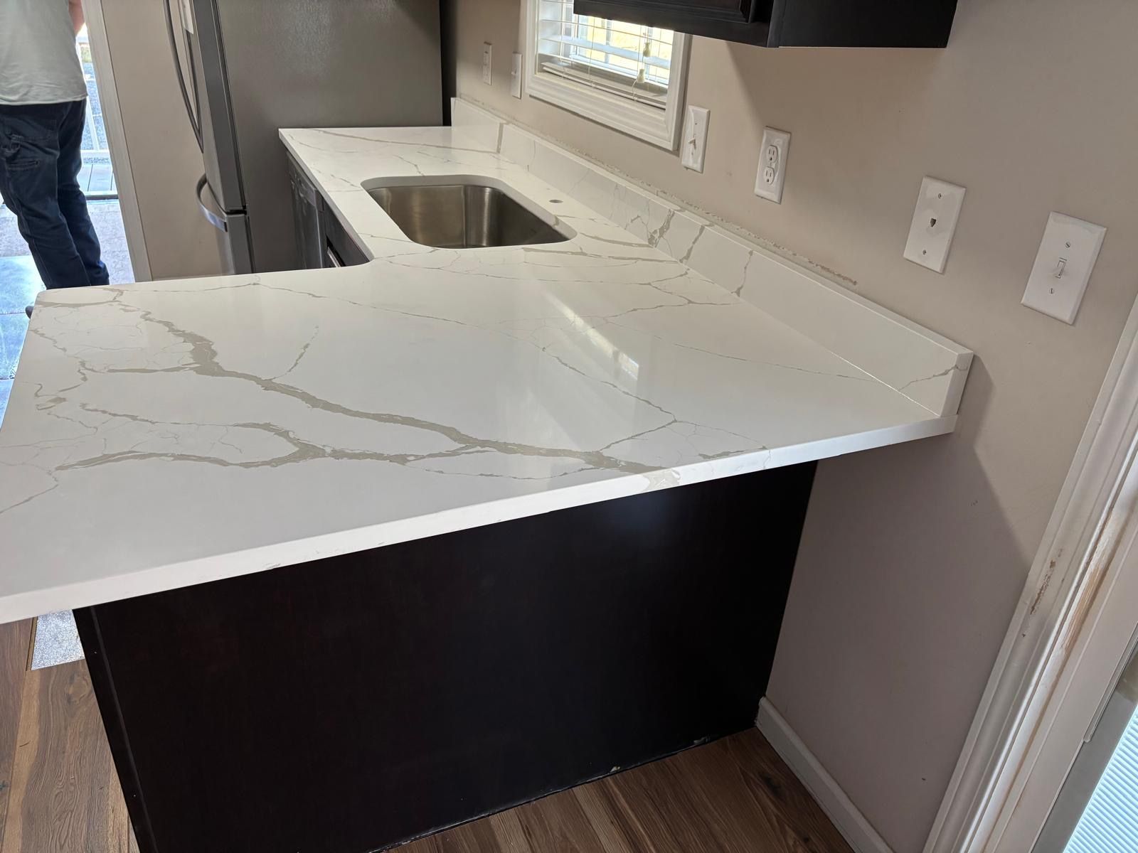 Kitchen counter with white quartz top, undermount sink, and dark cabinet base near a wall outlet.