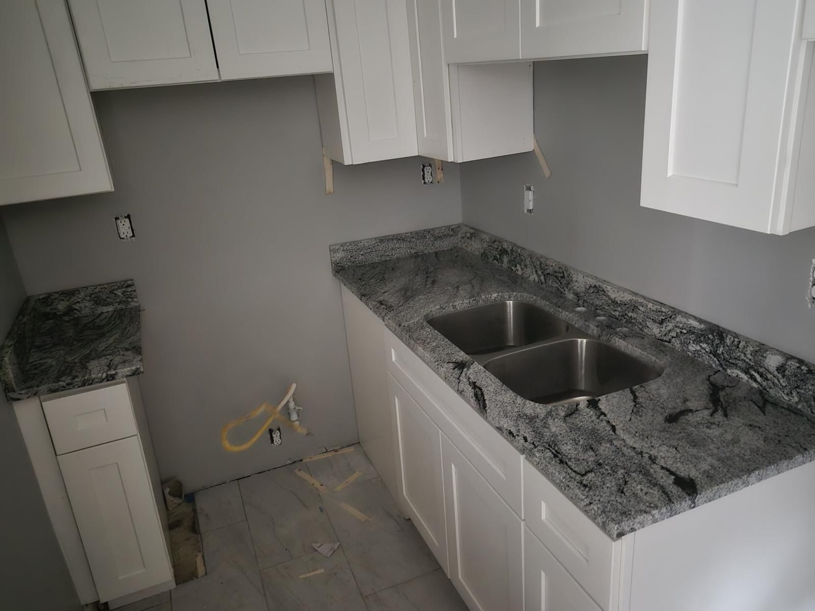 Unfinished kitchen with white cabinets, gray granite counters, and a double sink near exposed wiring.