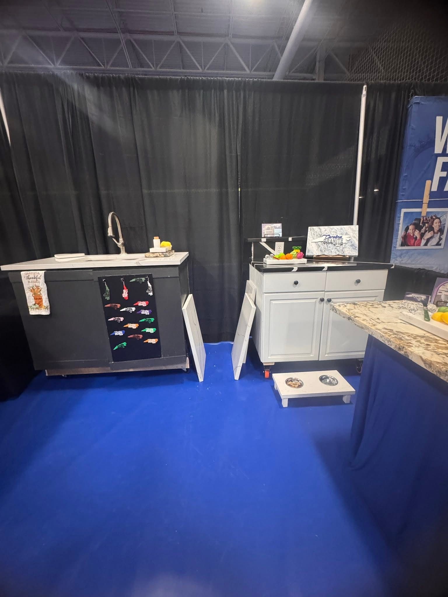 Trade show booth with black curtains, blue carpet, display tables, and a countertop under indoor lighting
