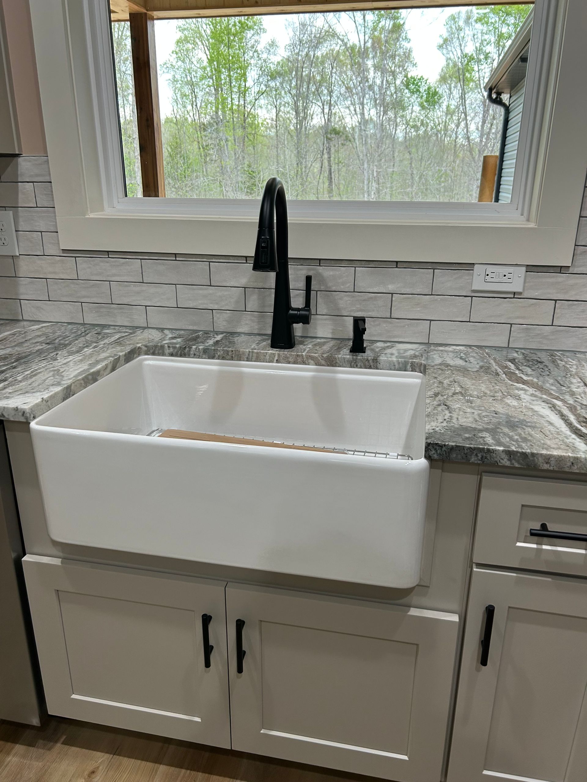 White farmhouse sink with black faucet in a kitchen, set against a window and gray stone backsplash.