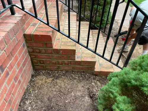 Brick steps and black metal railing beside a dirt patch and green shrub in a small outdoor yard