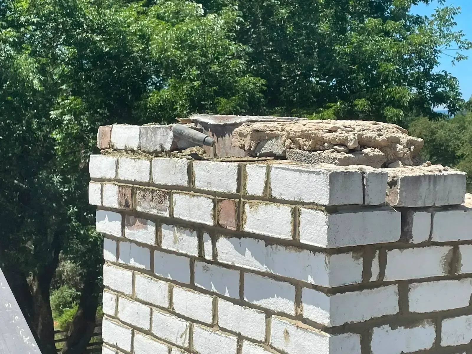 White brick chimney with crumbling top against leafy green trees outdoors