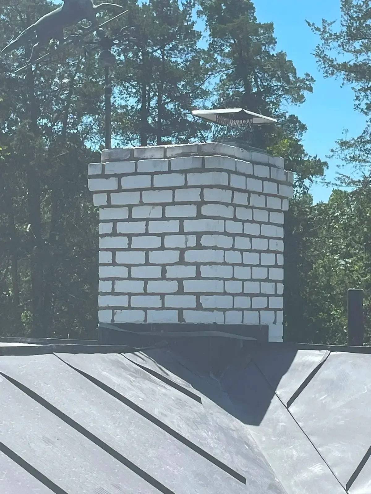 White brick chimney on a dark metal roof against trees and blue sky.
