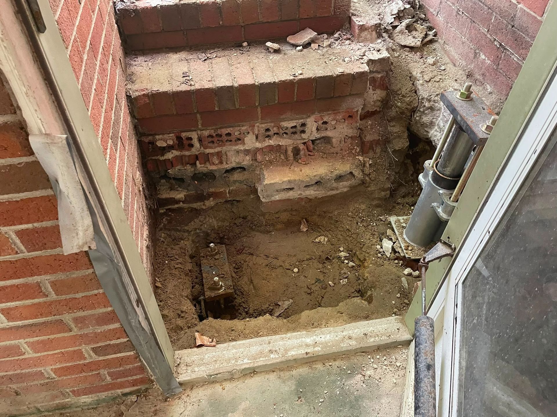 Exposed brick-lined pit beside a house foundation with dirt floor and broken concrete edge