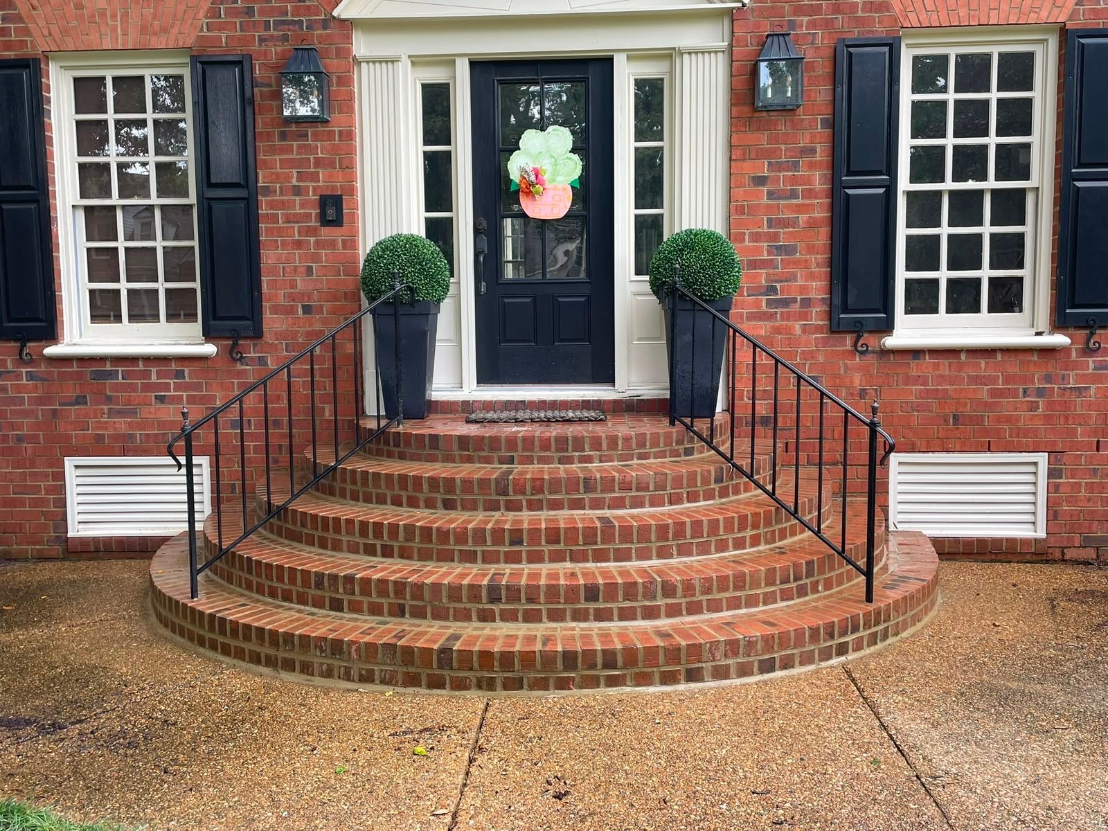 Brick house entrance with black door, white trim, and curved red brick steps lined with railings and potted shrubs
