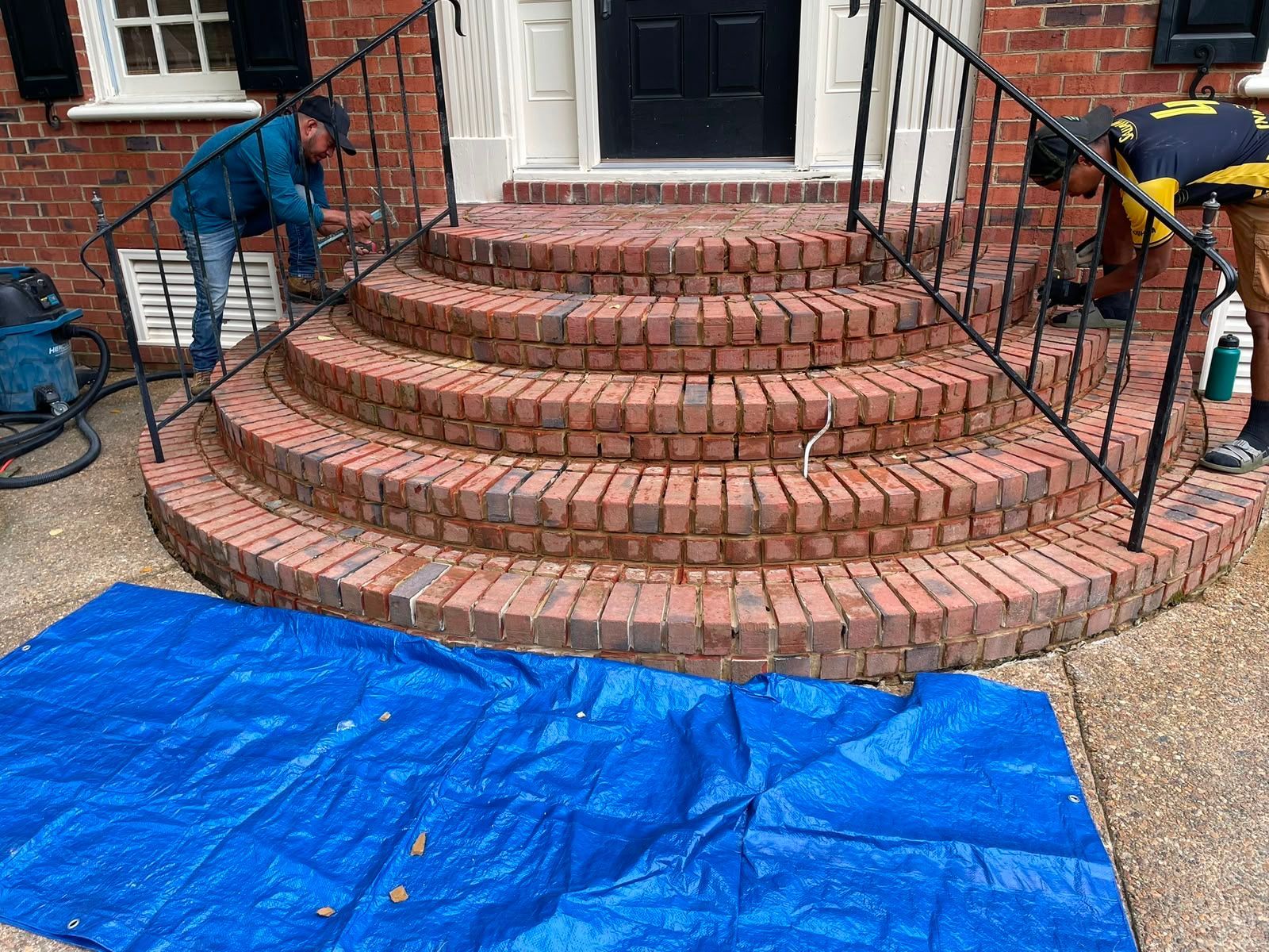 Workers repairing brick front steps with blue tarp and tools laid out on the ground