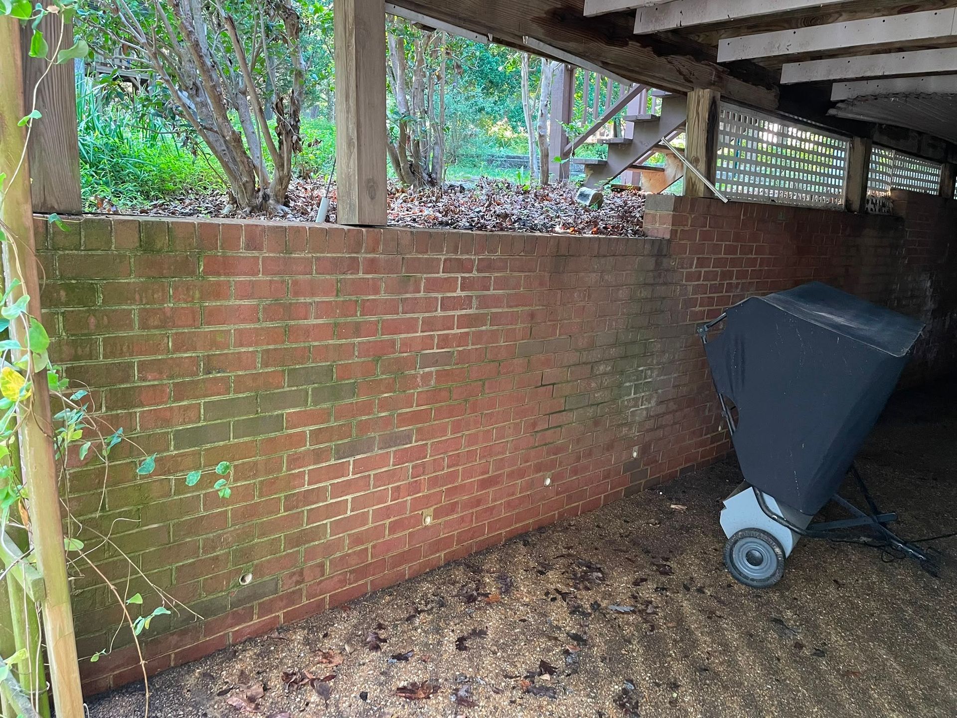 Brick wall under a covered patio with a large wheeled trash bin on dirt ground.