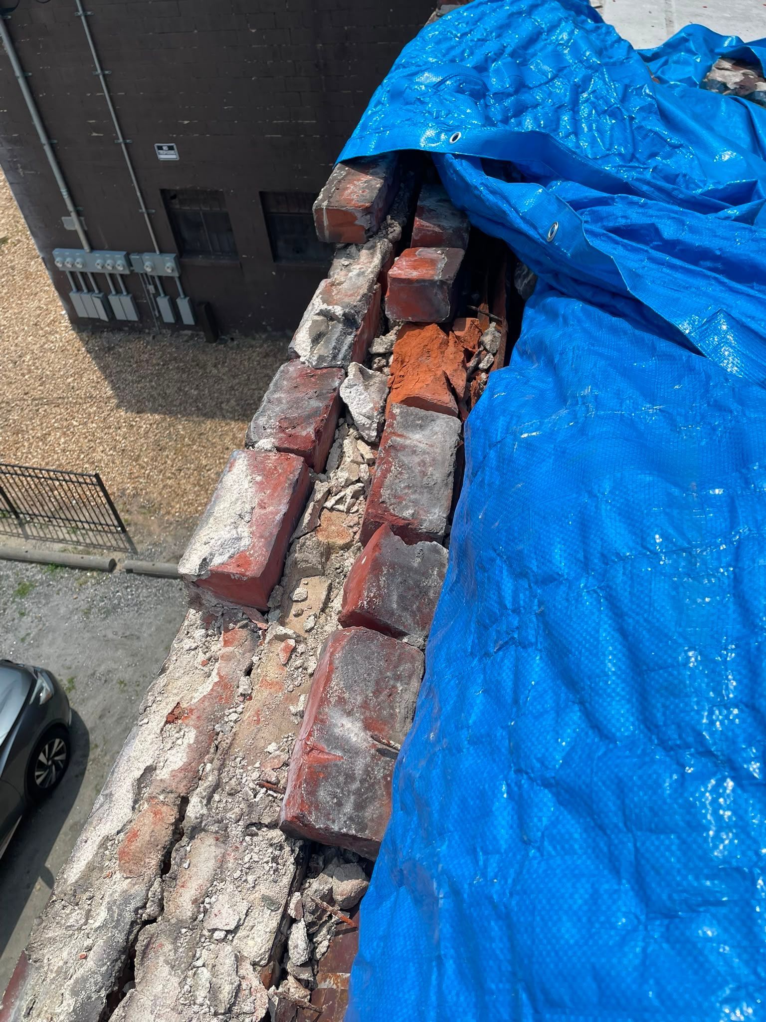Damaged brick rooftop parapet covered by a blue tarp beside a building.