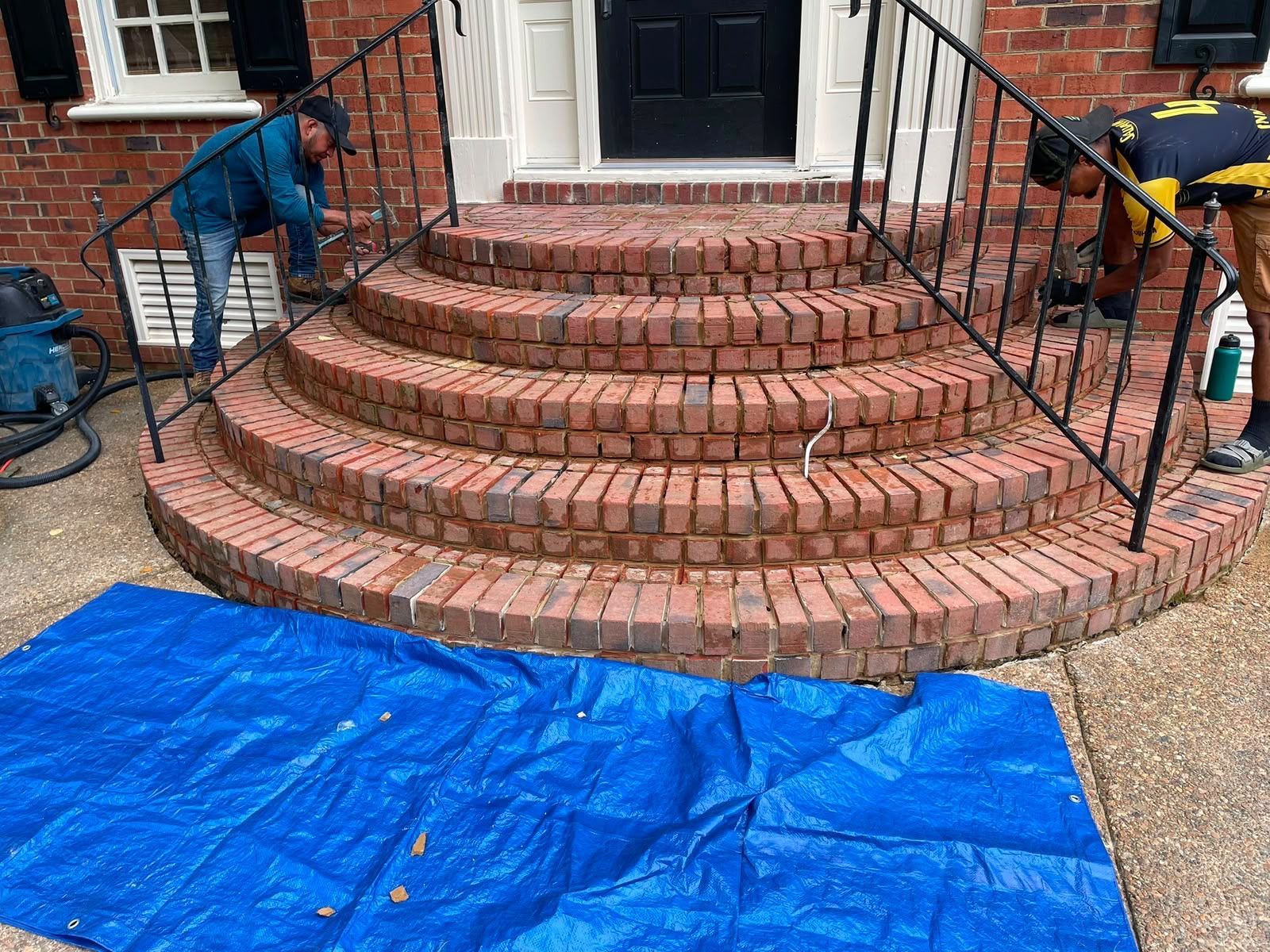 Two workers repairing brick front steps beside a blue tarp and black railings