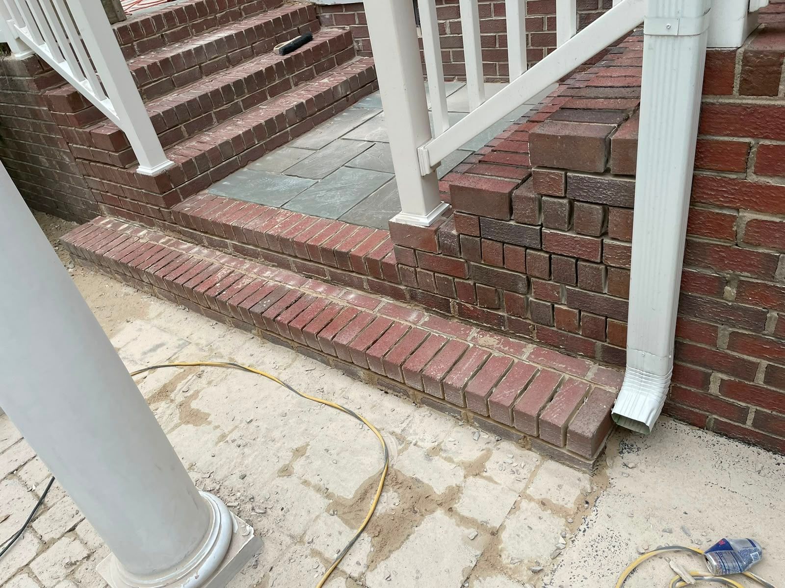 Brick porch steps with white railings and a brick wall, next to a cracked concrete walkway and downspout