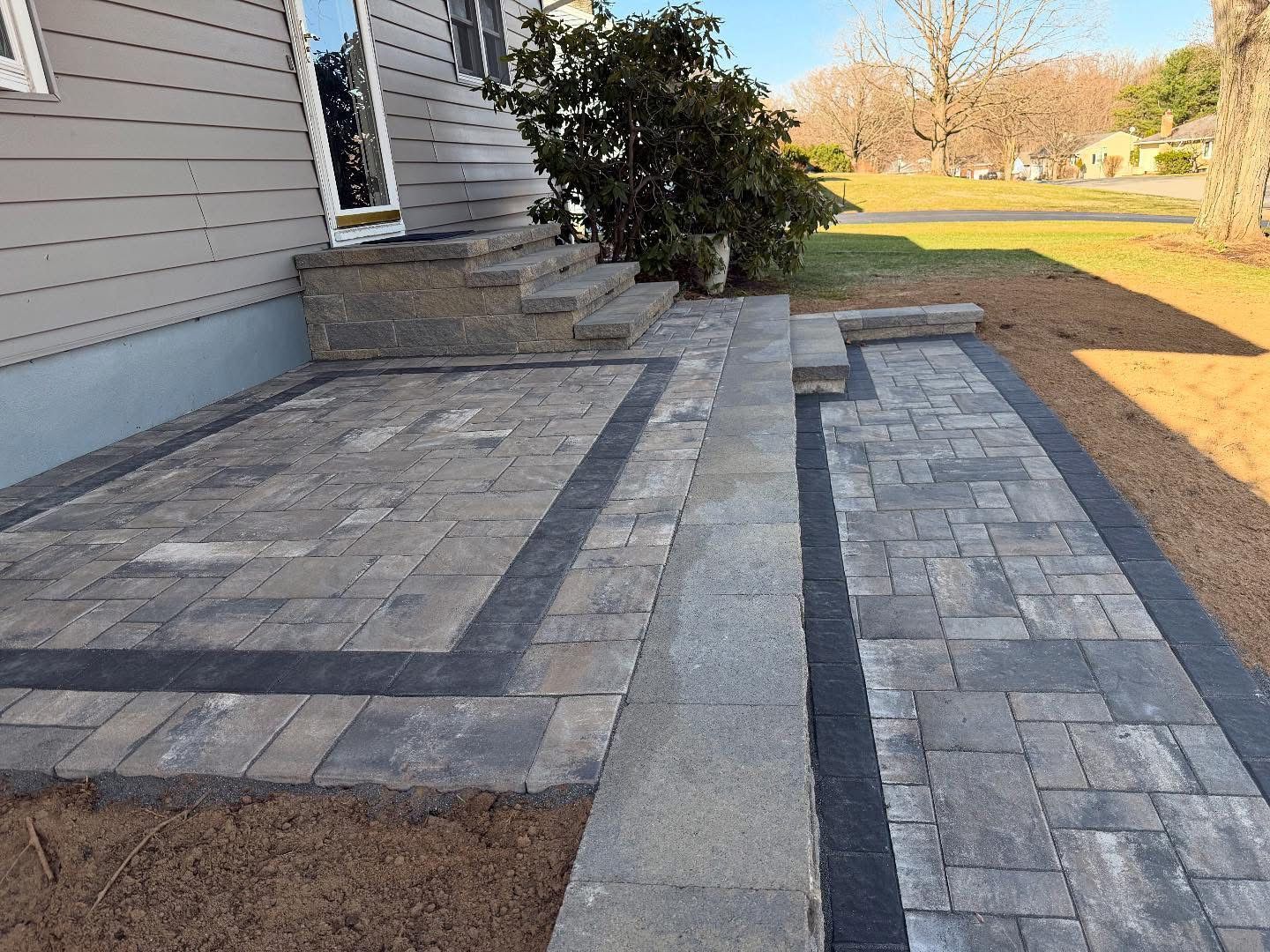 Stone patio and walkway beside a house, with gray pavers and a small step area leading to a grassy yard.