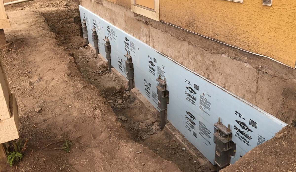 Foundation wall with blue waterproofing membrane beside a yellow building and dirt trench