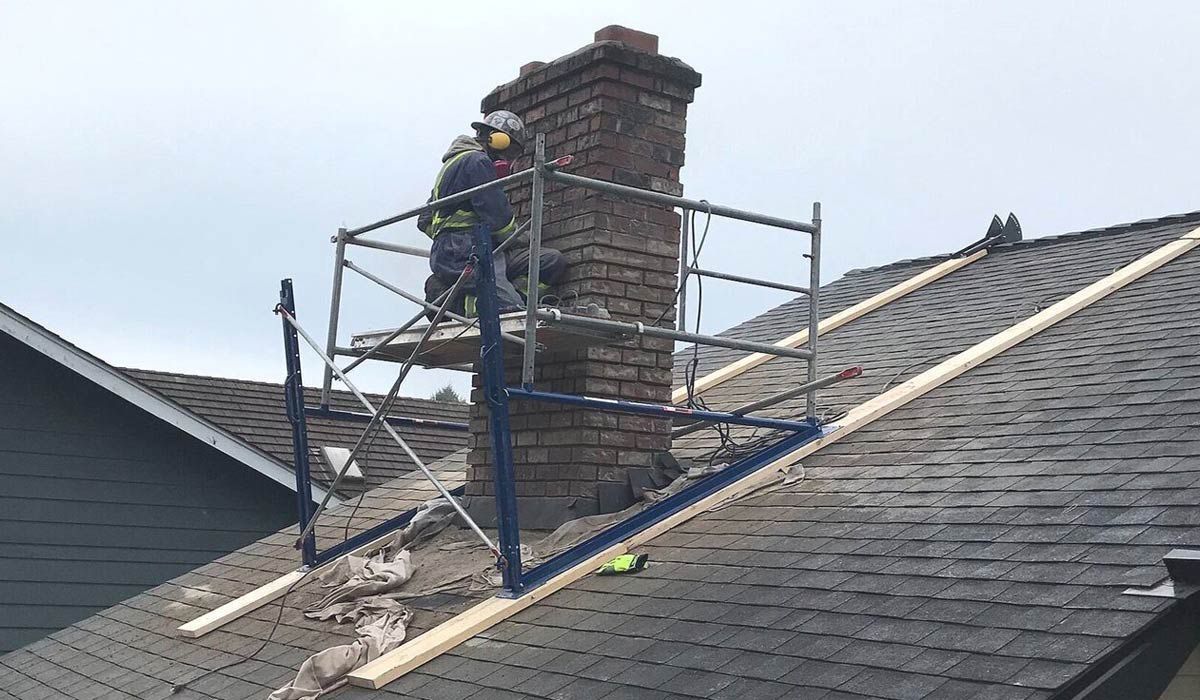 Worker on roof scaffolding beside a chimney, repairing shingles on a gray house roof.