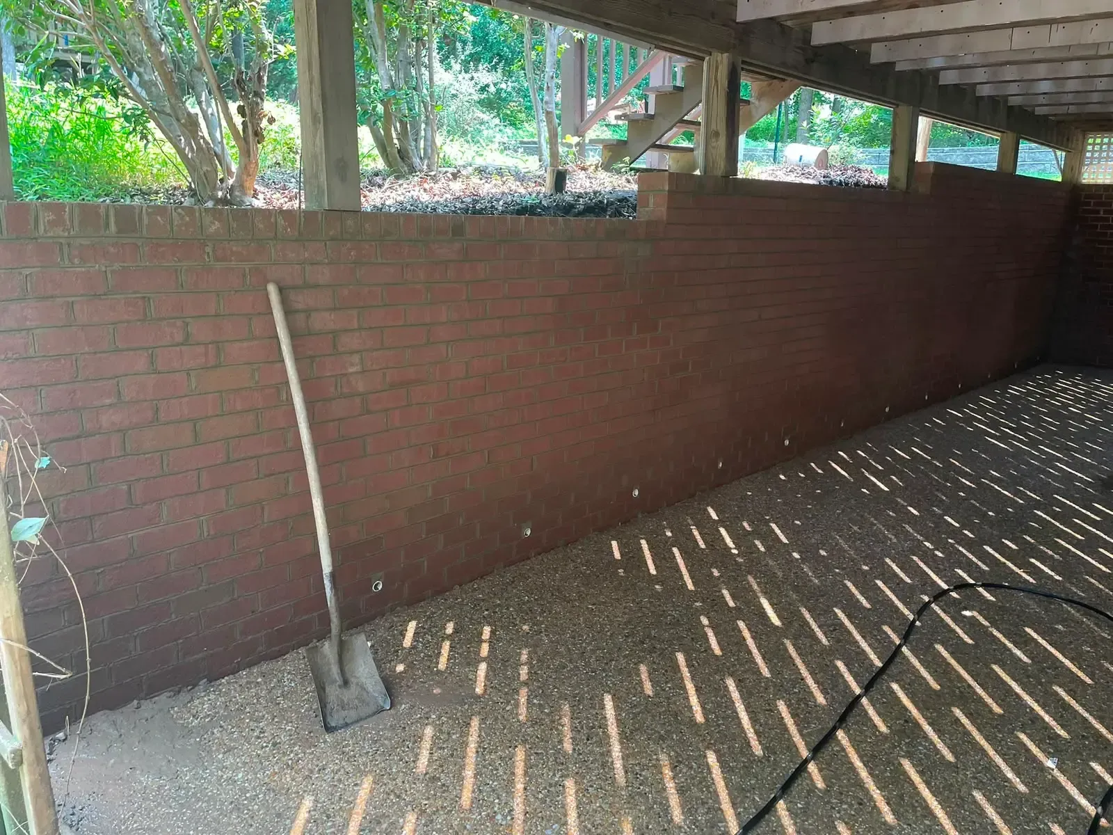 Brick retaining wall under a covered outdoor area, with dappled sunlight on the floor and a shovel leaning nearby