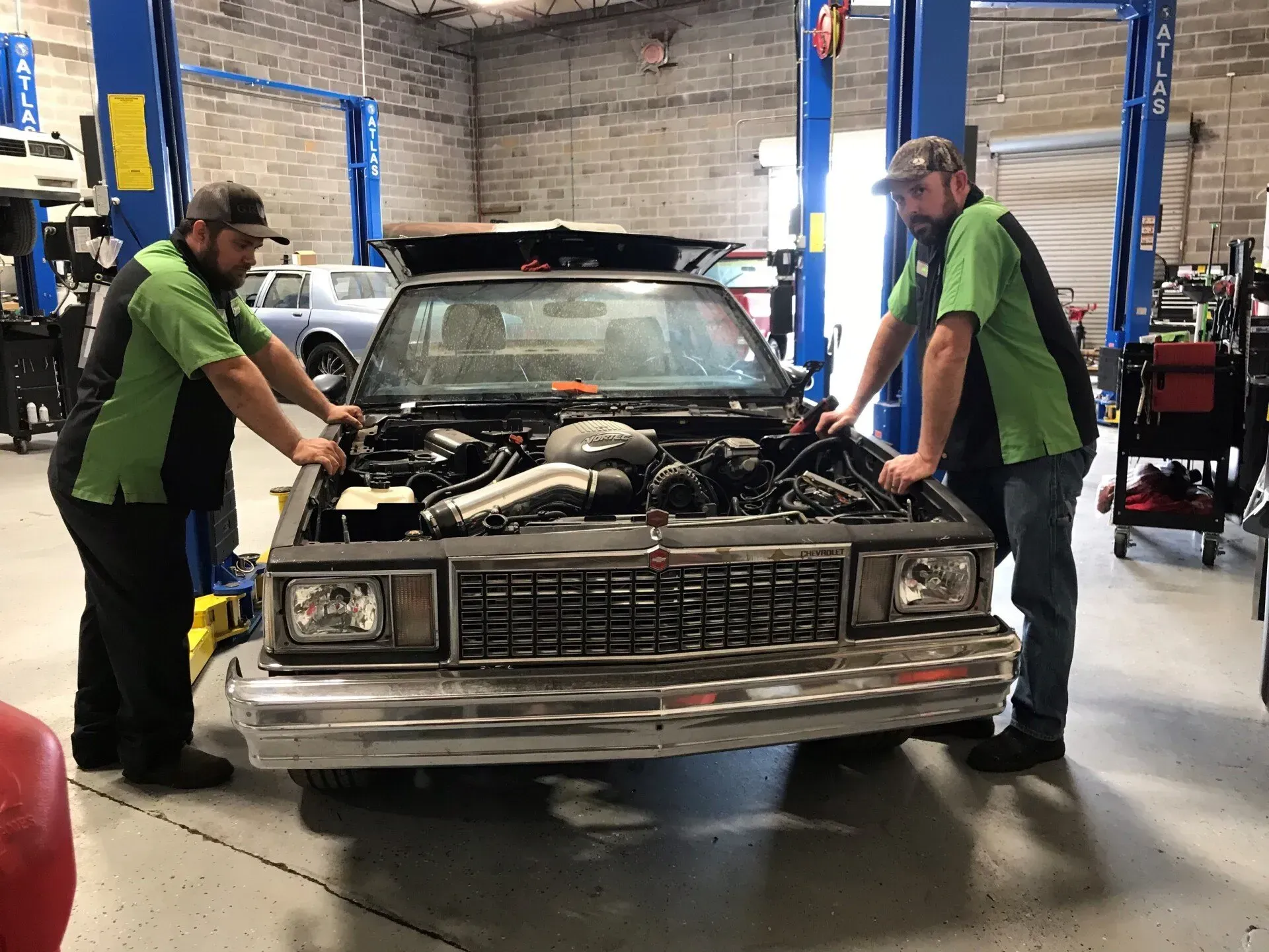 Two mechanics in green shirts examine a car engine in a garage.