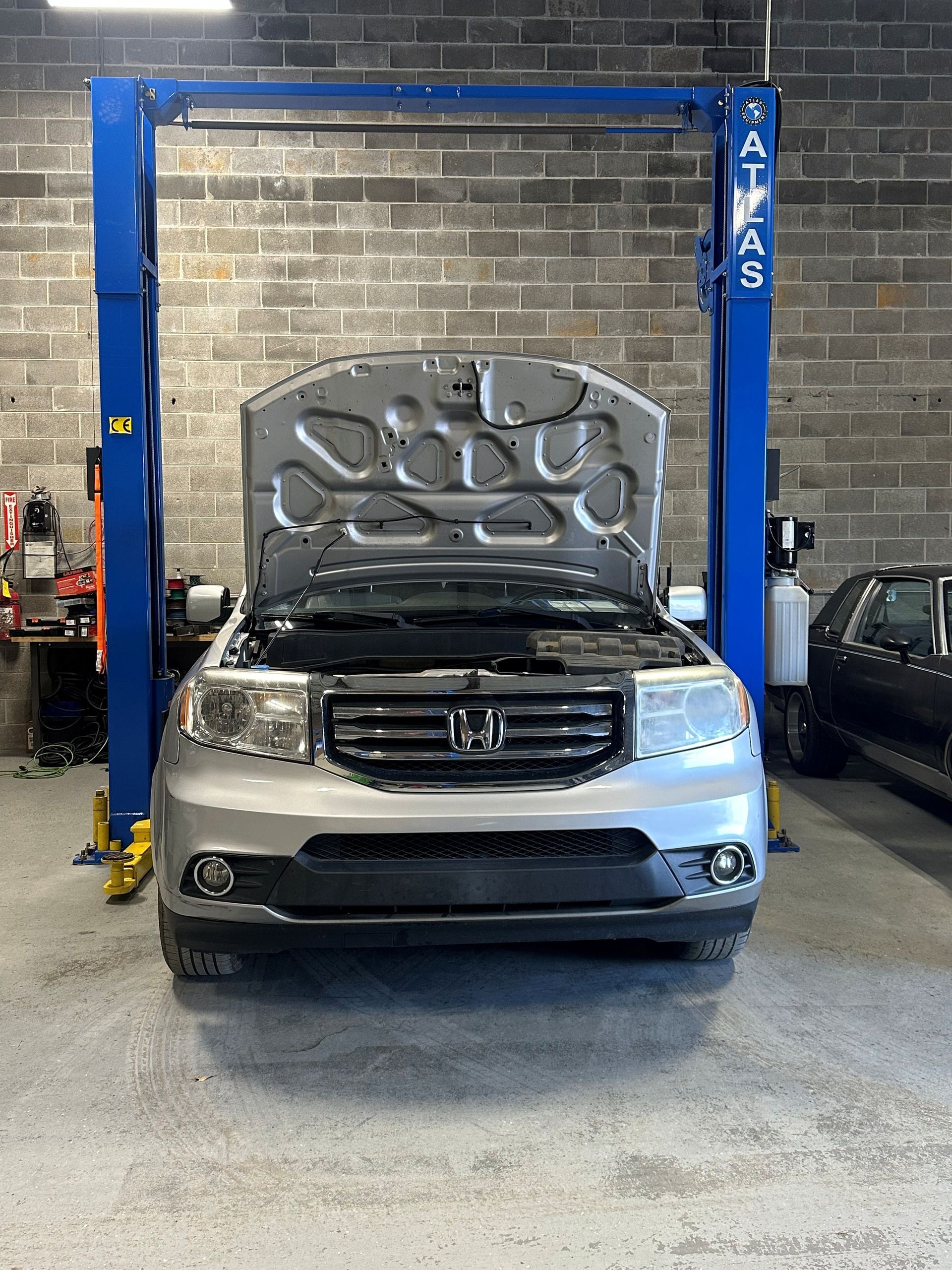 Silver Honda Pilot SUV on a lift with hood open inside a garage.