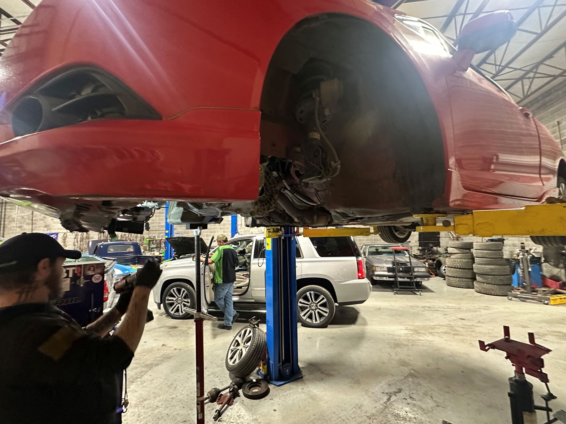 Mechanic working on a red car lifted in a garage; a silver SUV is in the background.