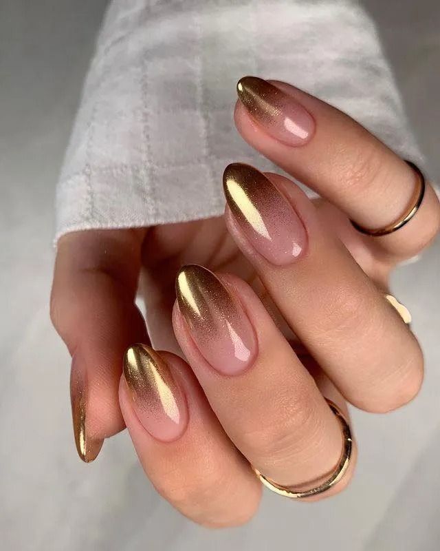Hand with oval-shaped nails featuring gold ombre tips, set against a pinkish-nude base, adorned with gold rings.