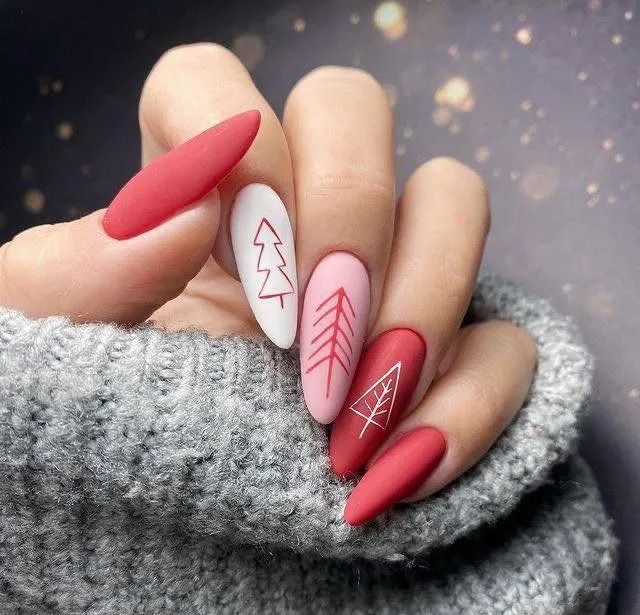 Hand with Christmas nail art: red, pink, and white nails with tree designs, next to a gray sweater.