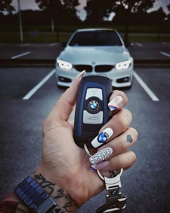 Hand holding a BMW key with a car in the background. The key has a BMW logo and a parking lot setting.