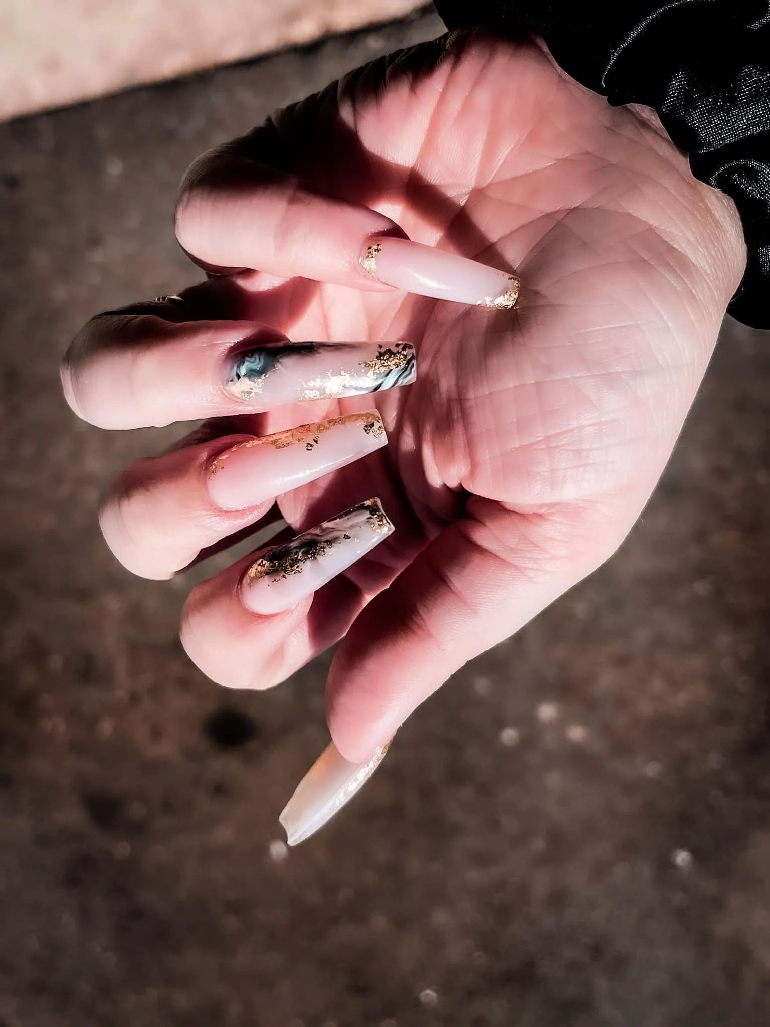 Hand with long, pale pink acrylic nails adorned with gold and iridescent accents.