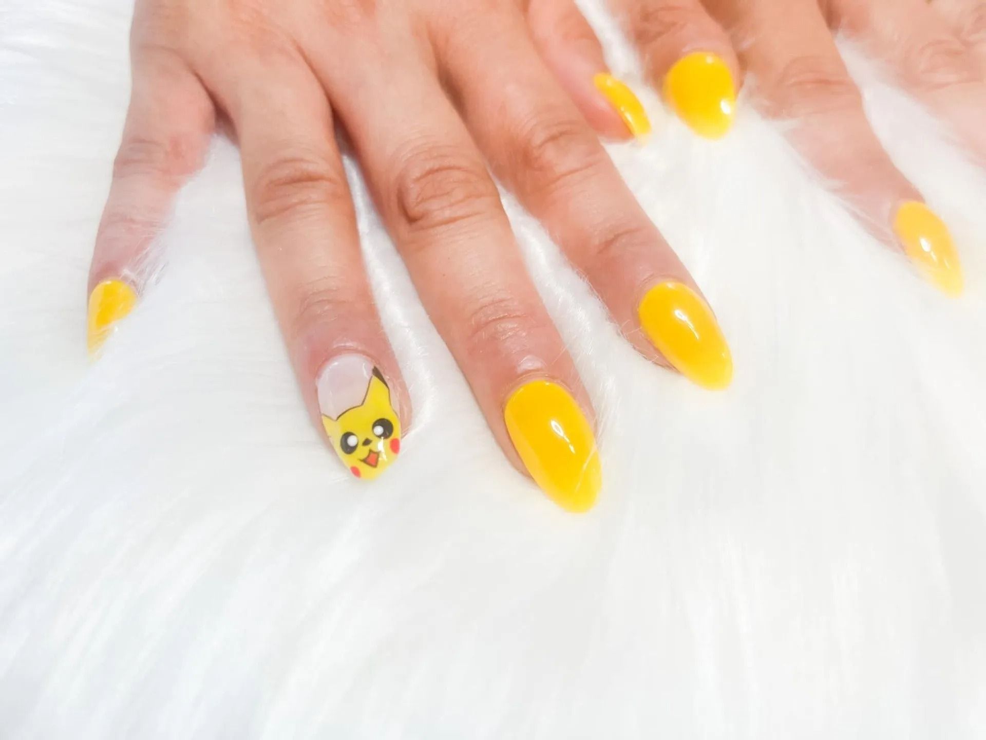 Hand with yellow almond-shaped nails, one featuring a Pikachu design, resting on a white, fluffy surface.