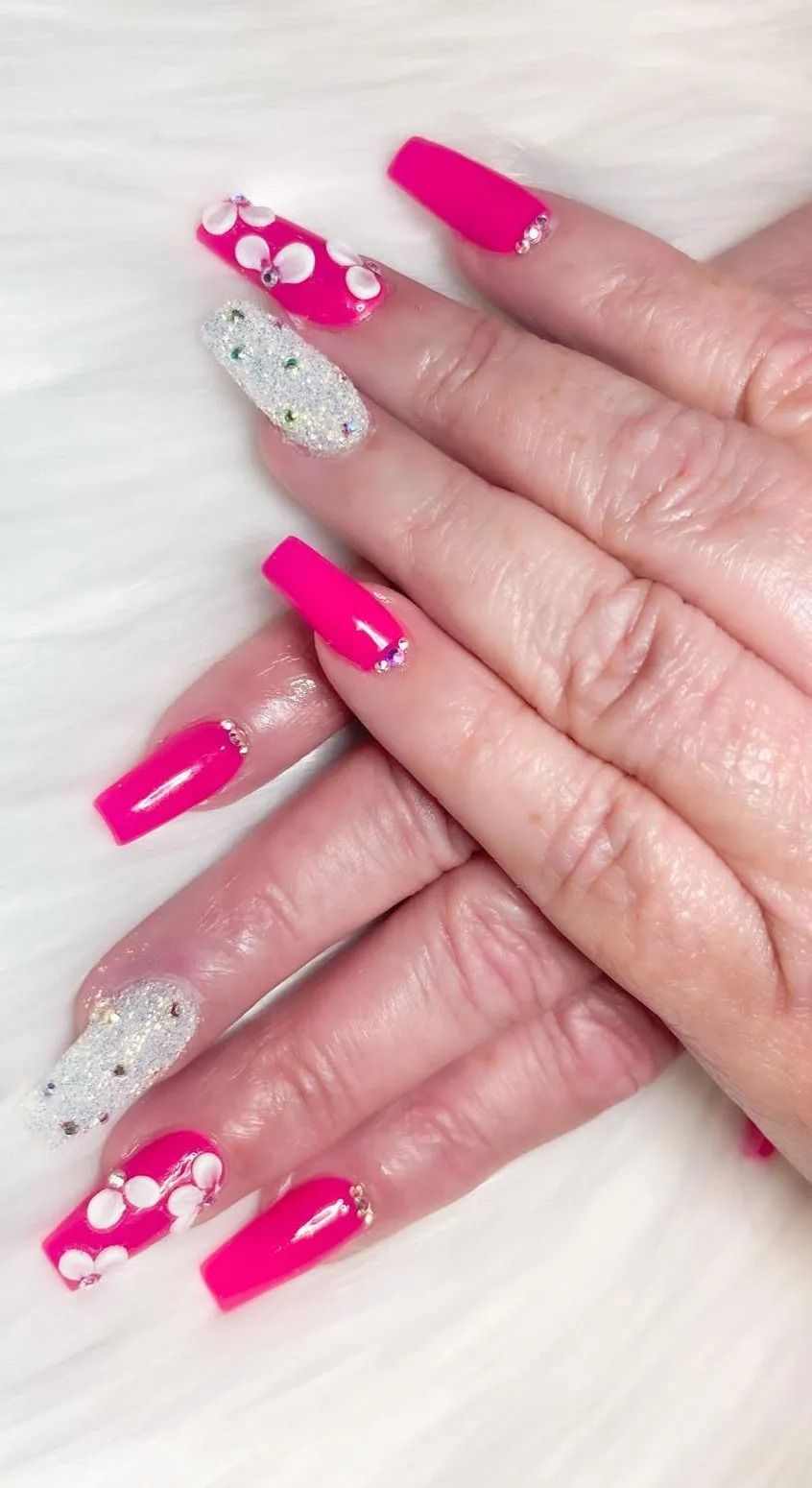 Hands with bright pink and glittery coffin-shaped nails, some decorated with white flower designs.