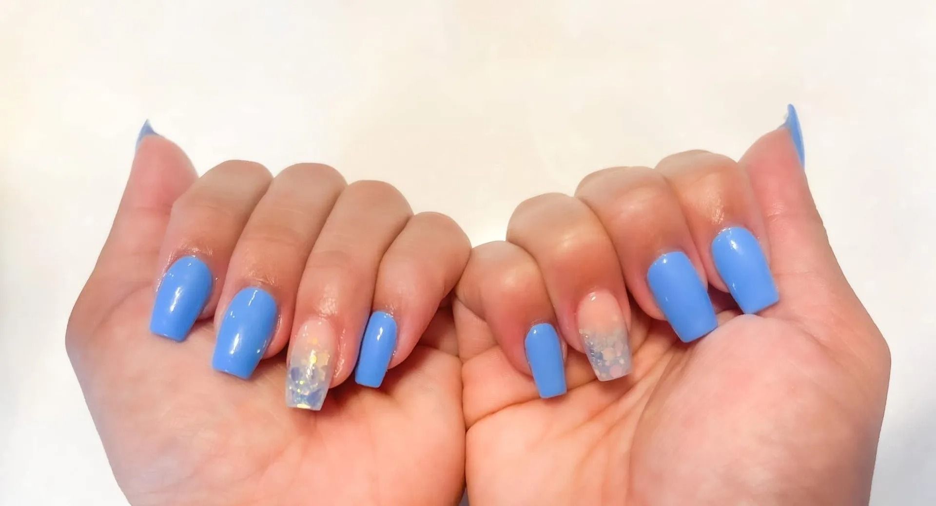 Hands with long, light blue, glossy fingernails and a clear glitter accent nail against a white background.