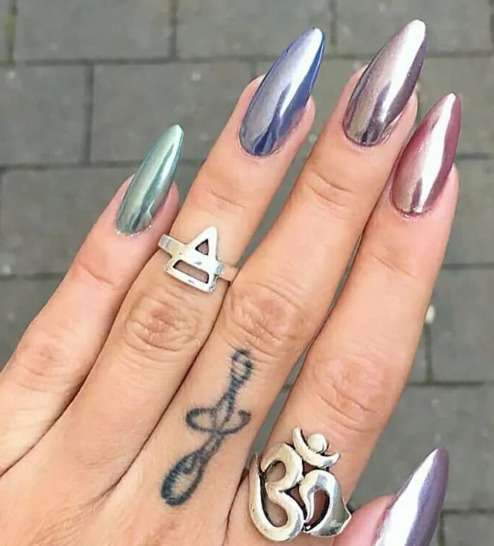 Hand with long, pointed, metallic-colored nails. Rings and finger tattoo visible.