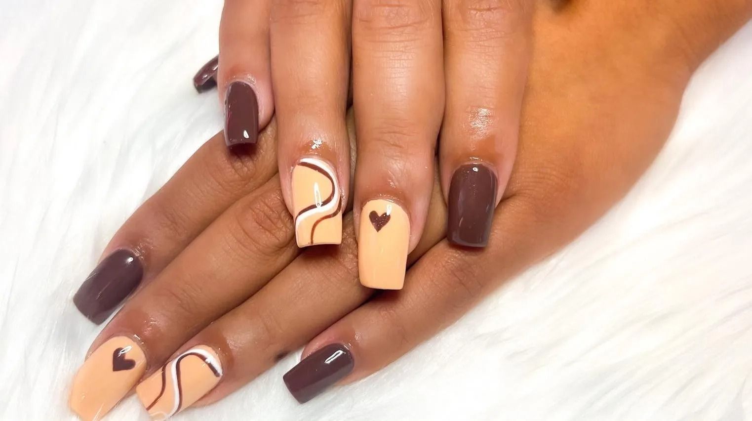 Hands with square-shaped nails in dark brown and nude colors, some with heart and swirl designs.