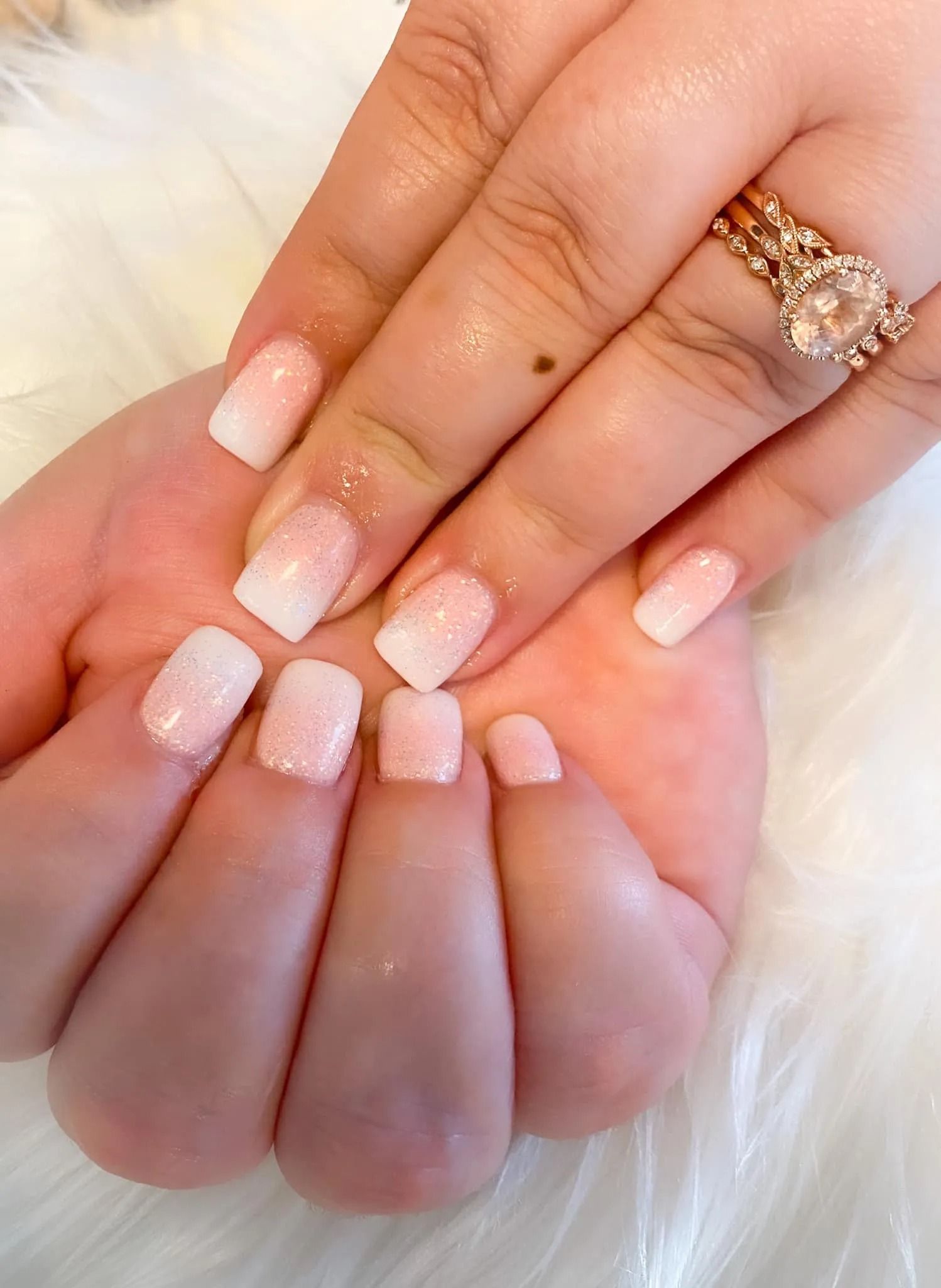 Hands with white and pink ombre nails, wearing a diamond ring, resting on a fluffy white surface.