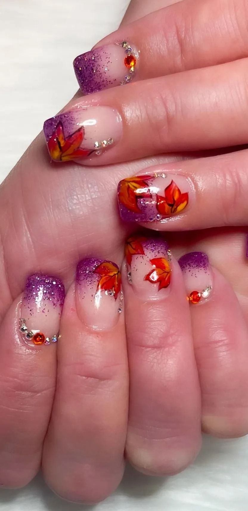 A hand with autumn-themed nail art. Featuring purple glitter, ombre, and leaf designs with rhinestones.