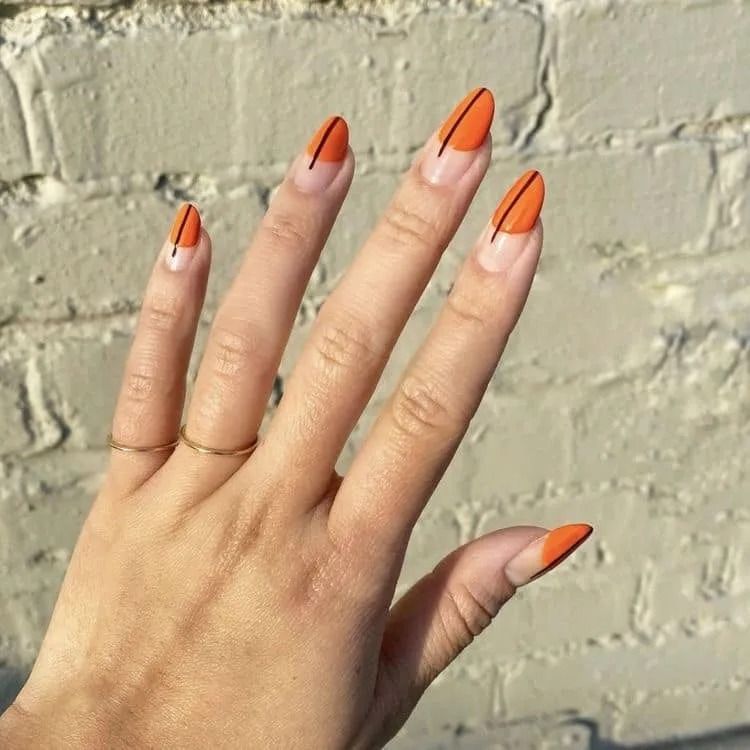 Hand with orange almond-shaped nails, black line design, gold ring, against a brick wall.