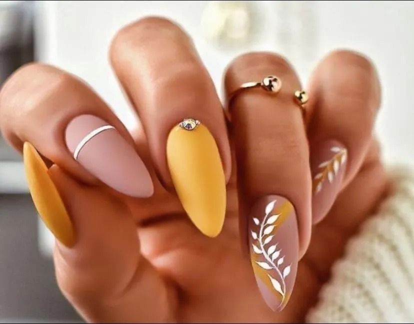 Hand with almond-shaped nails in matte yellow, mauve, and brown, featuring designs with lines, gems, and leaves.
