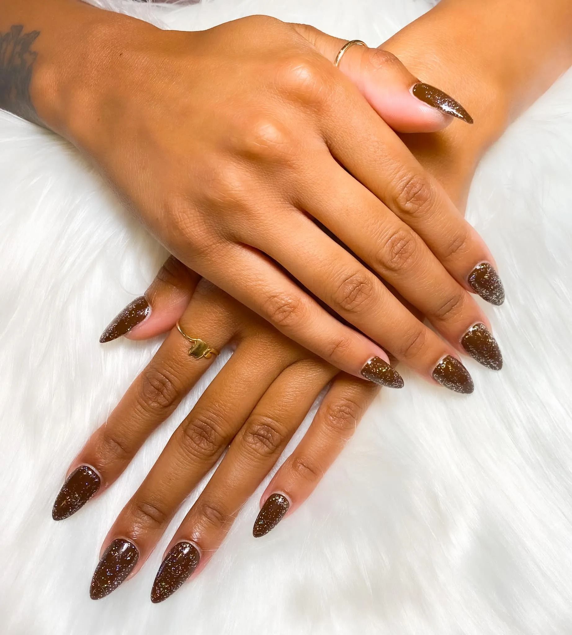 Hands with long, pointed, dark brown sparkly nails, crossed on a white fluffy surface.