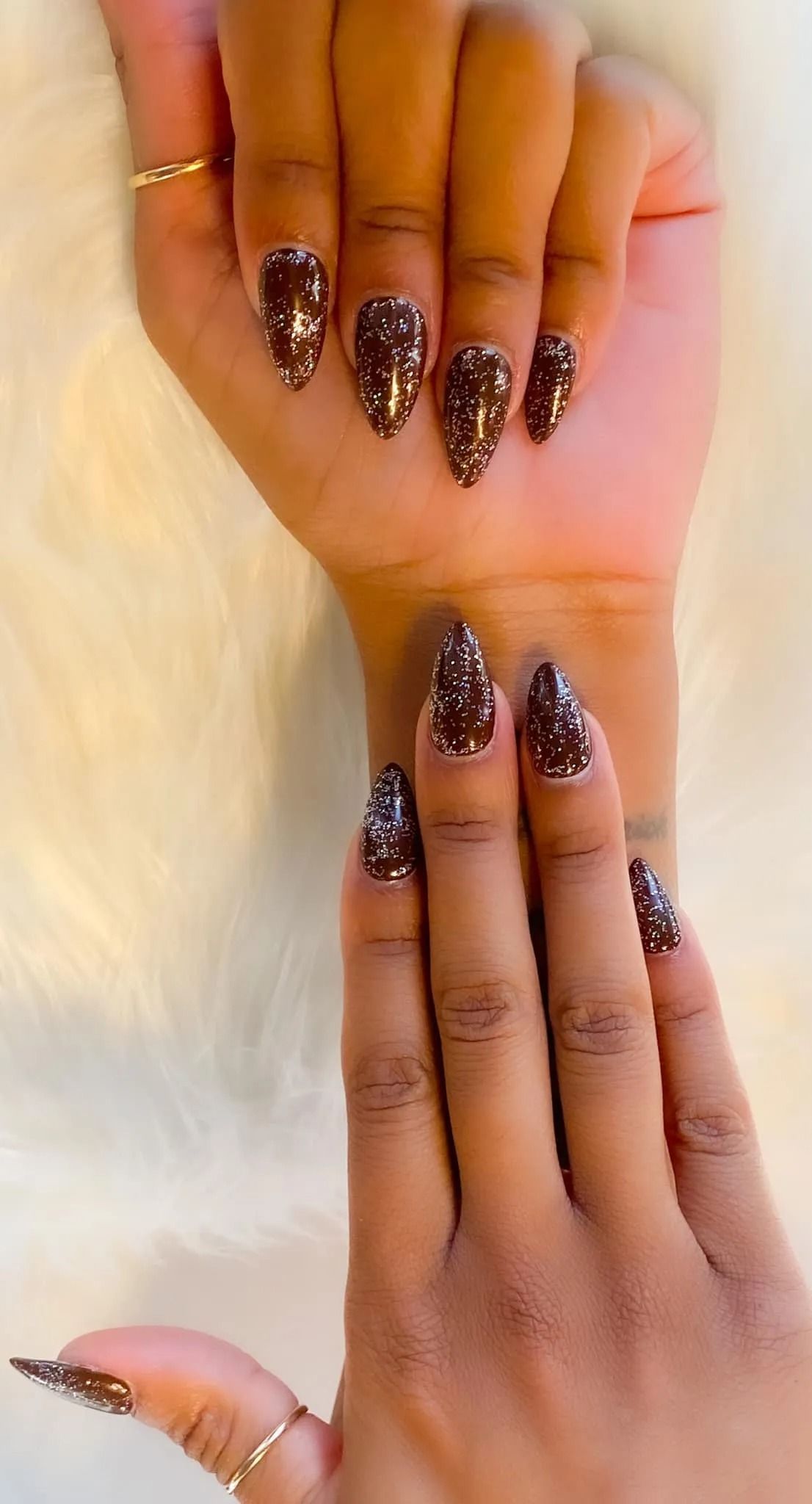 Hands with stiletto-shaped nails; dark brown base with gold glitter. One hand over the other, both resting on white fur.