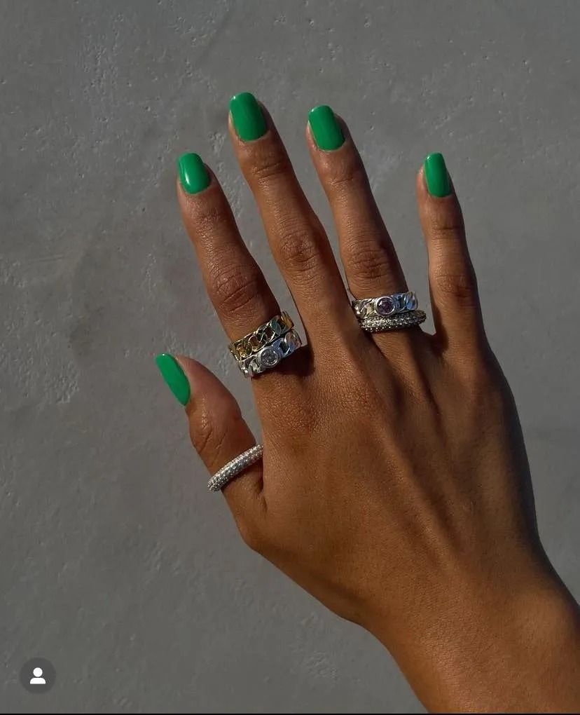 Hand with green nails, wearing multiple silver rings, against a neutral-colored background.