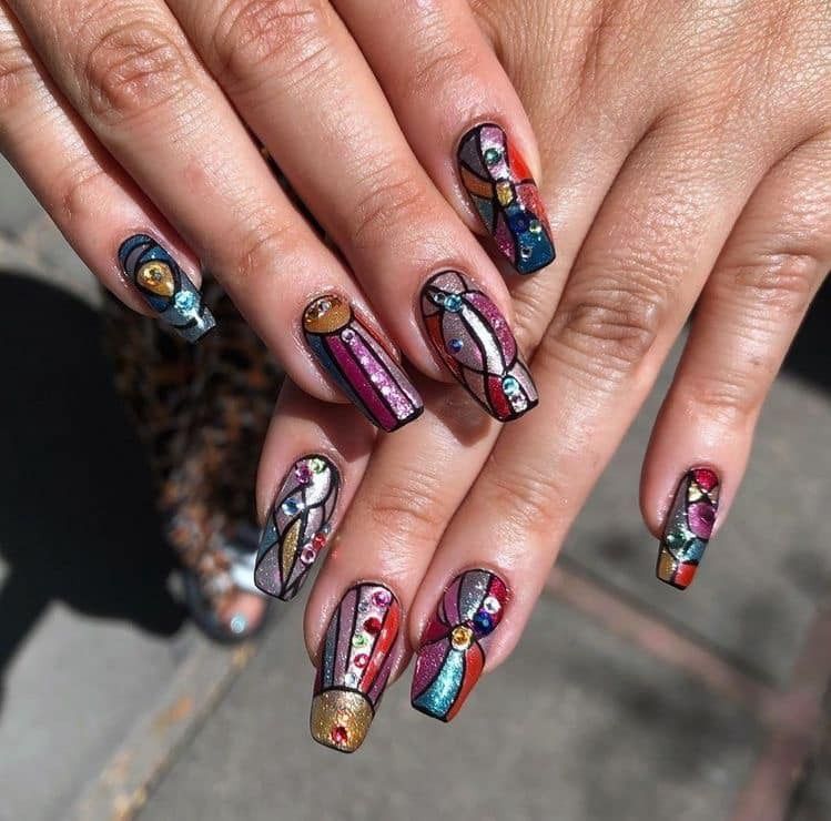 Hands with colorful, stained glass-inspired nail art. Geometric shapes in various colors with metallic outlines and gems.