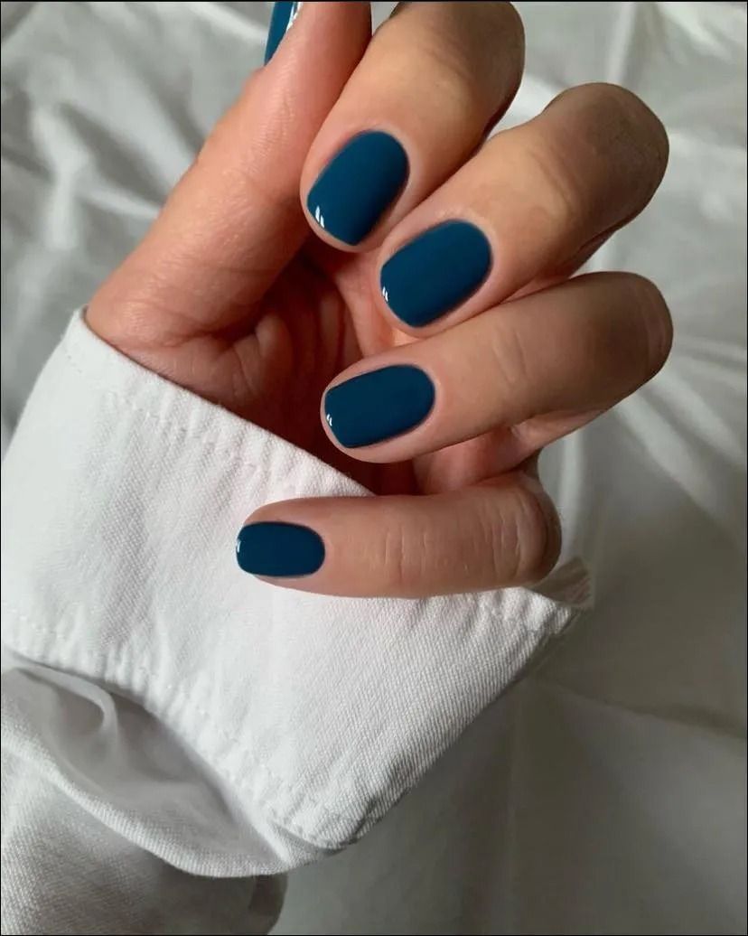 Hand with oval-shaped nails painted a dark teal color, resting on a white fabric.