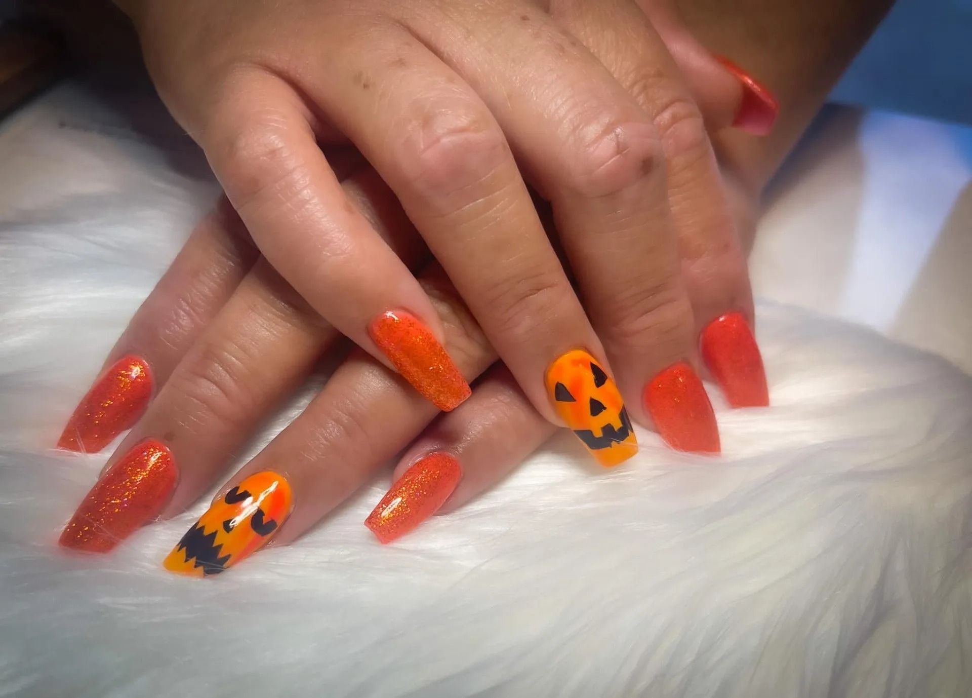 Orange Halloween nail art: long, coffin-shaped nails, some with glitter, and two with painted jack-o'-lantern faces.