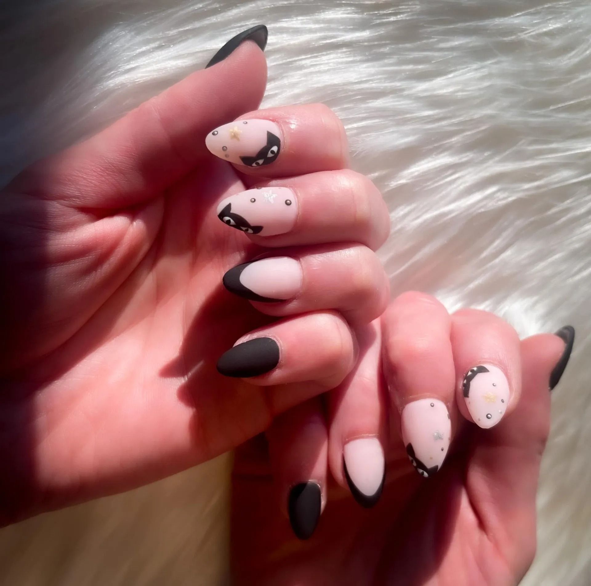 Hands with oval-shaped matte black and light pink nails. Some feature crescent moon and cat designs.