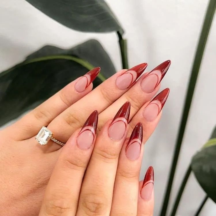Hand with long, pointed nails in red and nude. A ring is visible. Green plant background.