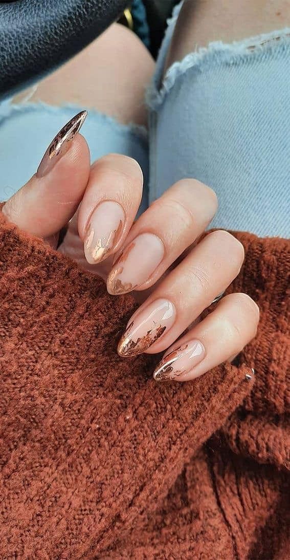 Hand with almond-shaped nails, some nude with gold glitter tips, resting on a brown sweater.