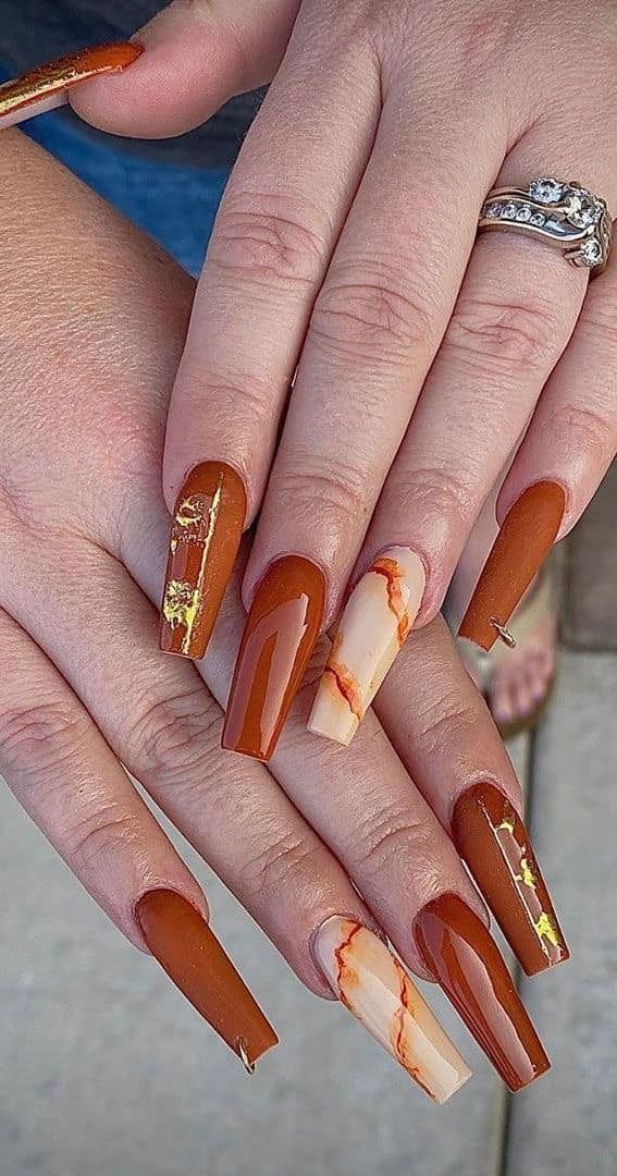 Hands with long coffin-shaped nails painted in shades of brown with marble and gold accents, and a silver ring.