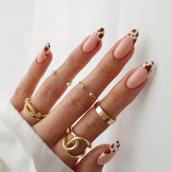 Hand with almond-shaped nails with cow print tips, wearing gold rings.