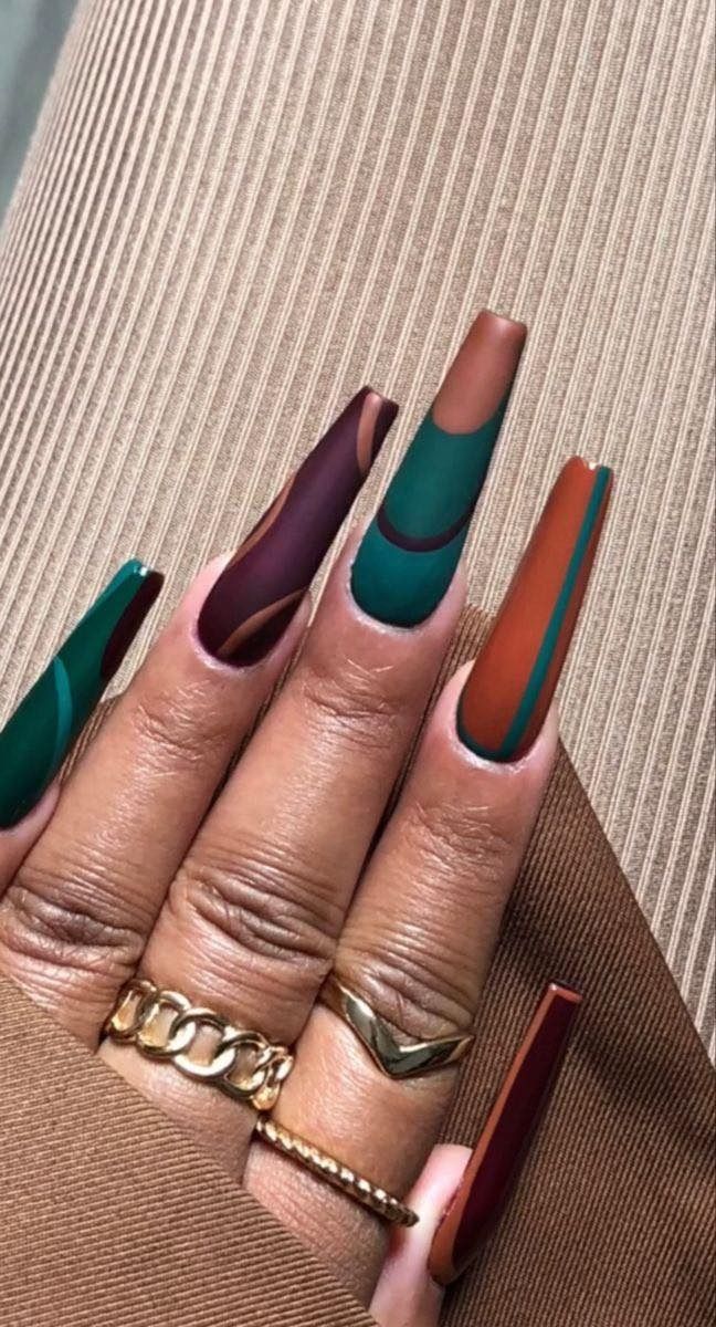 Long, coffin-shaped nails with abstract designs in shades of green, brown, and maroon. The hand is adorned with gold rings.